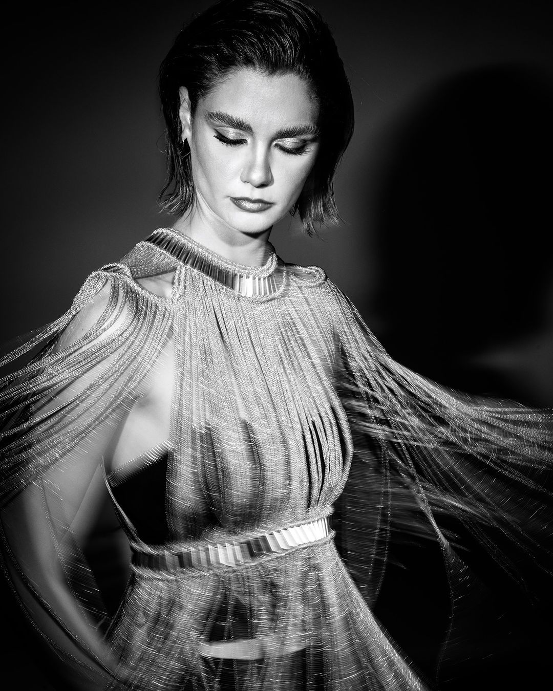 Black and white portrait of an adult woman in an elegant fringed dress, looking down against a dark background.