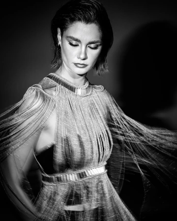 Black and white portrait of an adult woman in an elegant fringed dress, looking down against a dark background.