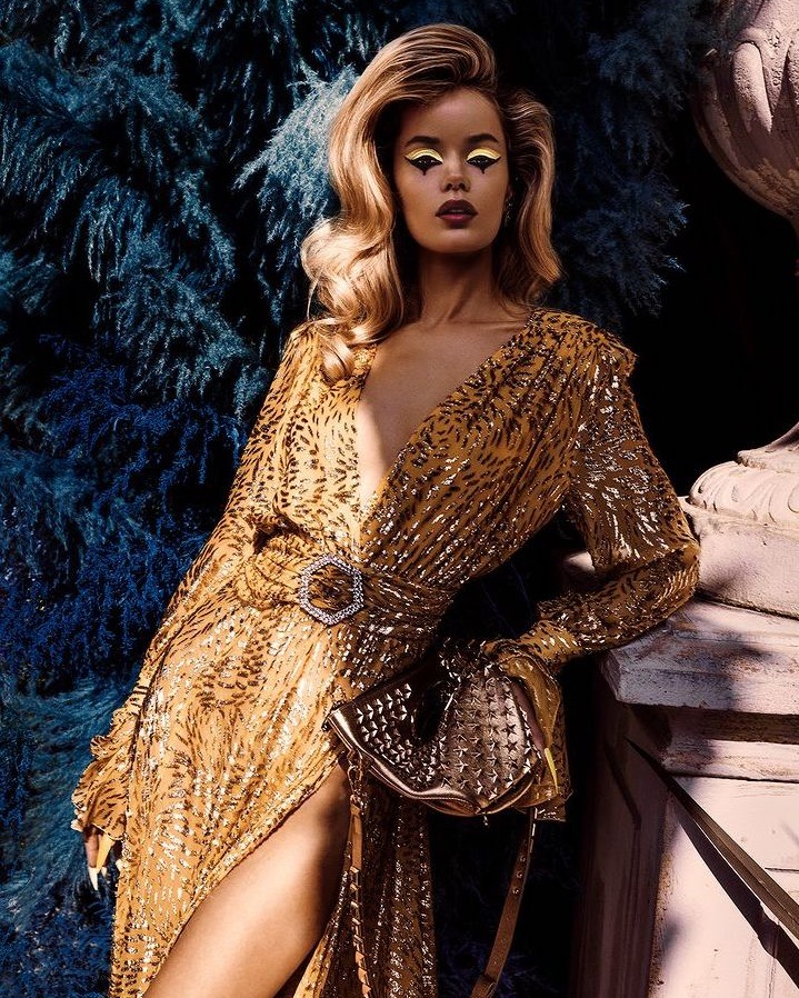 Adult female in animal-print gown with geometric eye makeup standing near a decorative blue evergreen bush and marble balustrade.