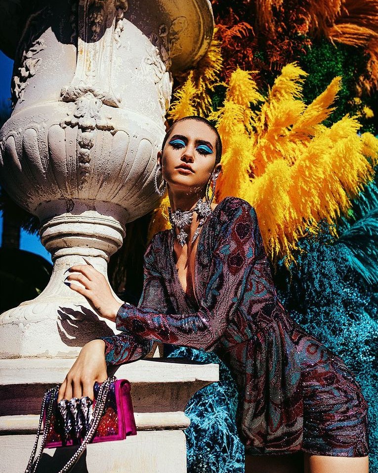 Elegant adult female poses near a carved stone urn with colorful floral arrangement, modelling jewelry and patterned dress, outdoors in daytime.