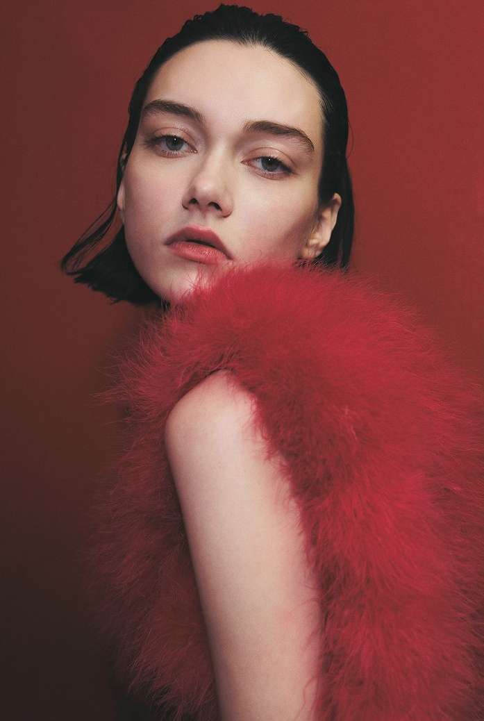 Portrait of a young woman with dark hair wearing a red, feathery garment against a red backdrop, gazing at the camera in soft light.