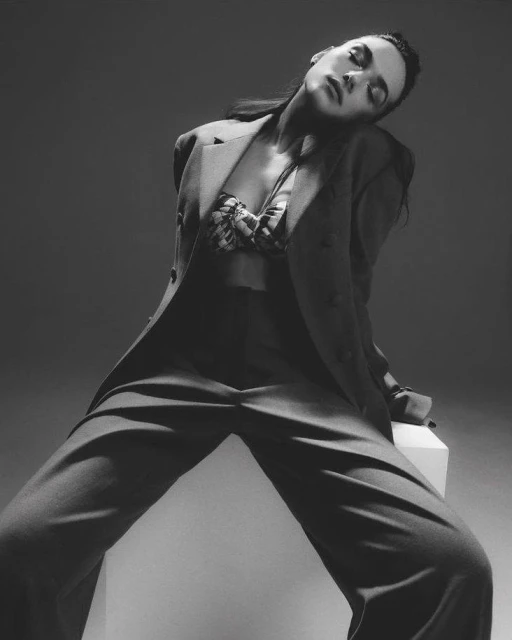 Monochrome portrait of a stylish young woman in a suit and bra top, leaning back against a white block in a photo studio setting.