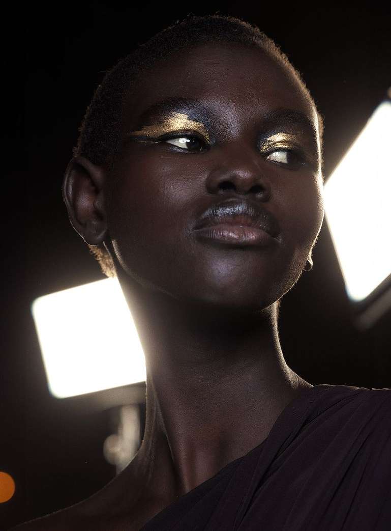 Close-up portrait of an adult Black woman with short hair and metallic gold eye makeup, wearing dark brown clothing against a dark background.