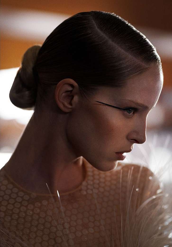Close-up portrait of an adult female with slicked back dark hair and dramatic eye makeup, facing left, wearing a textured beige dress.