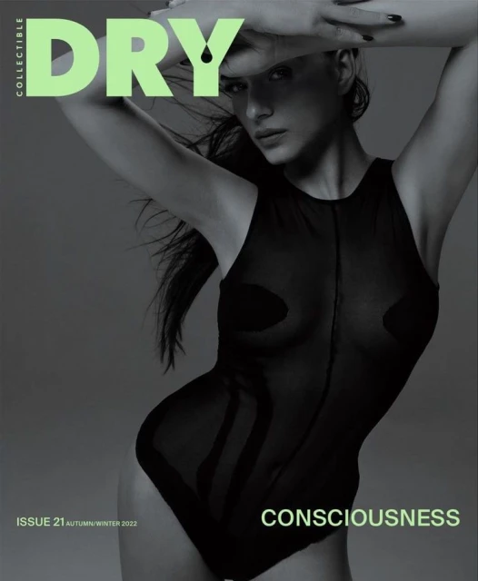 Fashion magazine cover features a female model in a sheer bodysuit, posing with hands above her head against a soft grey background.