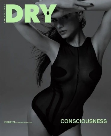 Fashion magazine cover features a female model in a sheer bodysuit, posing with hands above her head against a soft grey background.