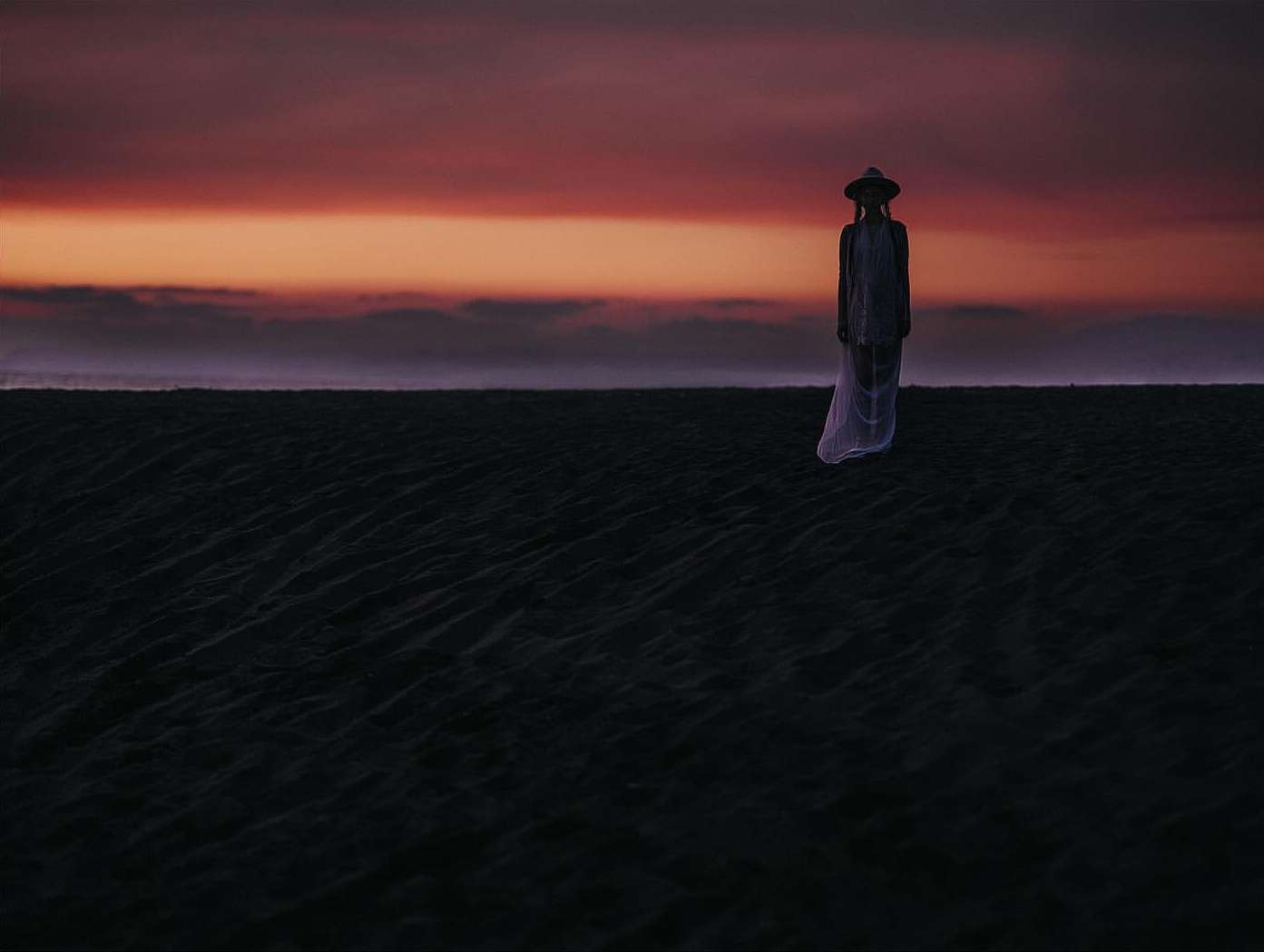 Silhouette of a woman wearing a hat and dress stands on a dark beach under a sunset sky with pink, orange, and purple hues.