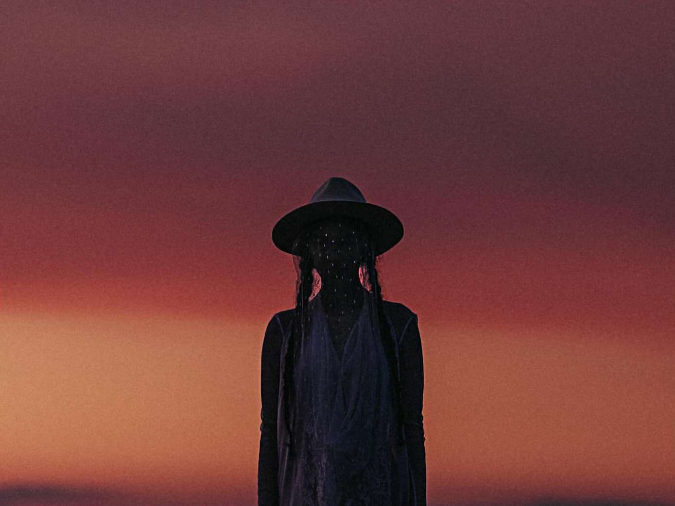 Silhouette of a person wearing a hat against a gradient orange and purple sky at sunset, creating a serene and atmospheric outdoor scene.