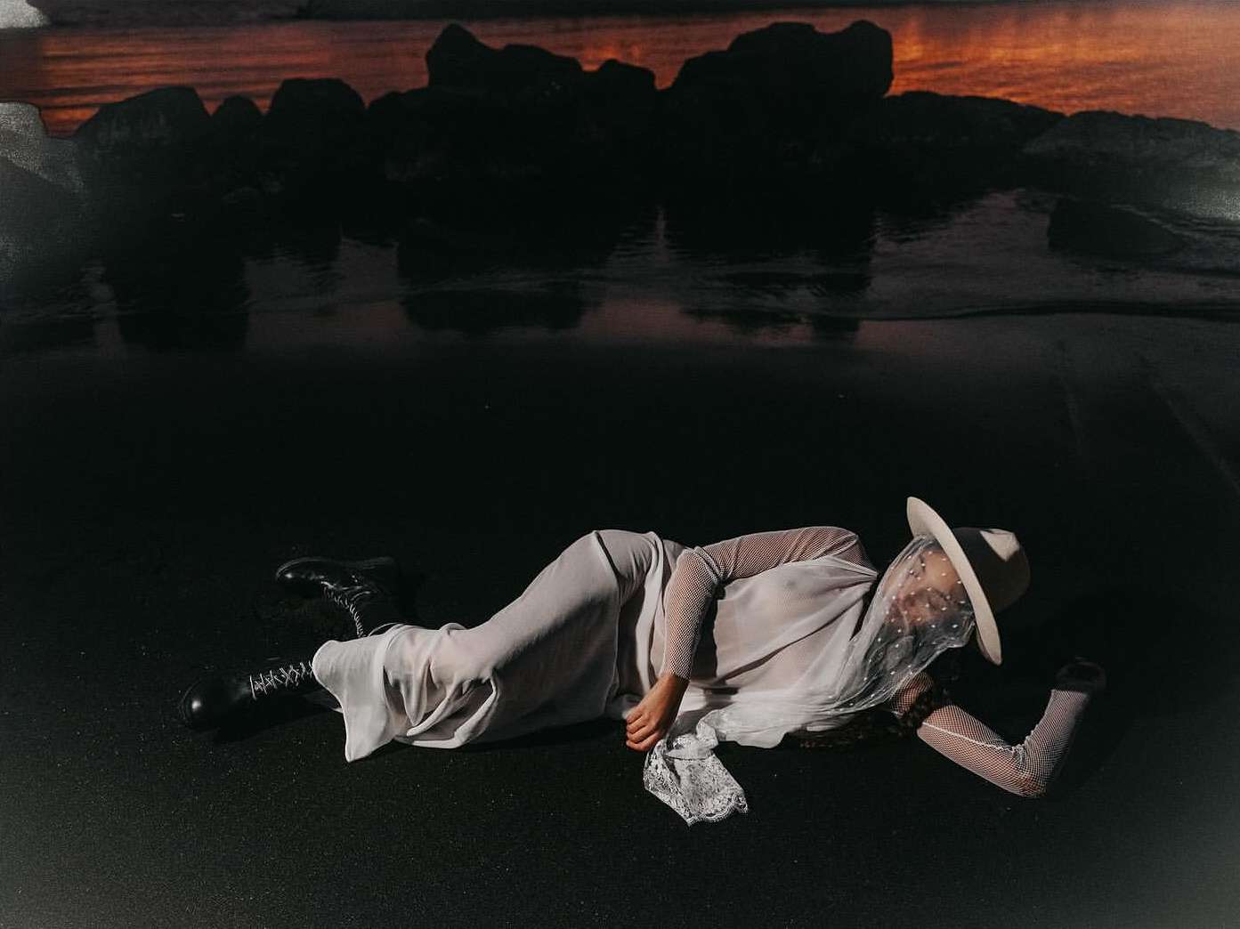 Woman in white dress and hat lies on black sand beach with rocky ocean backdrop, photographed with soft light and shallow depth of field.