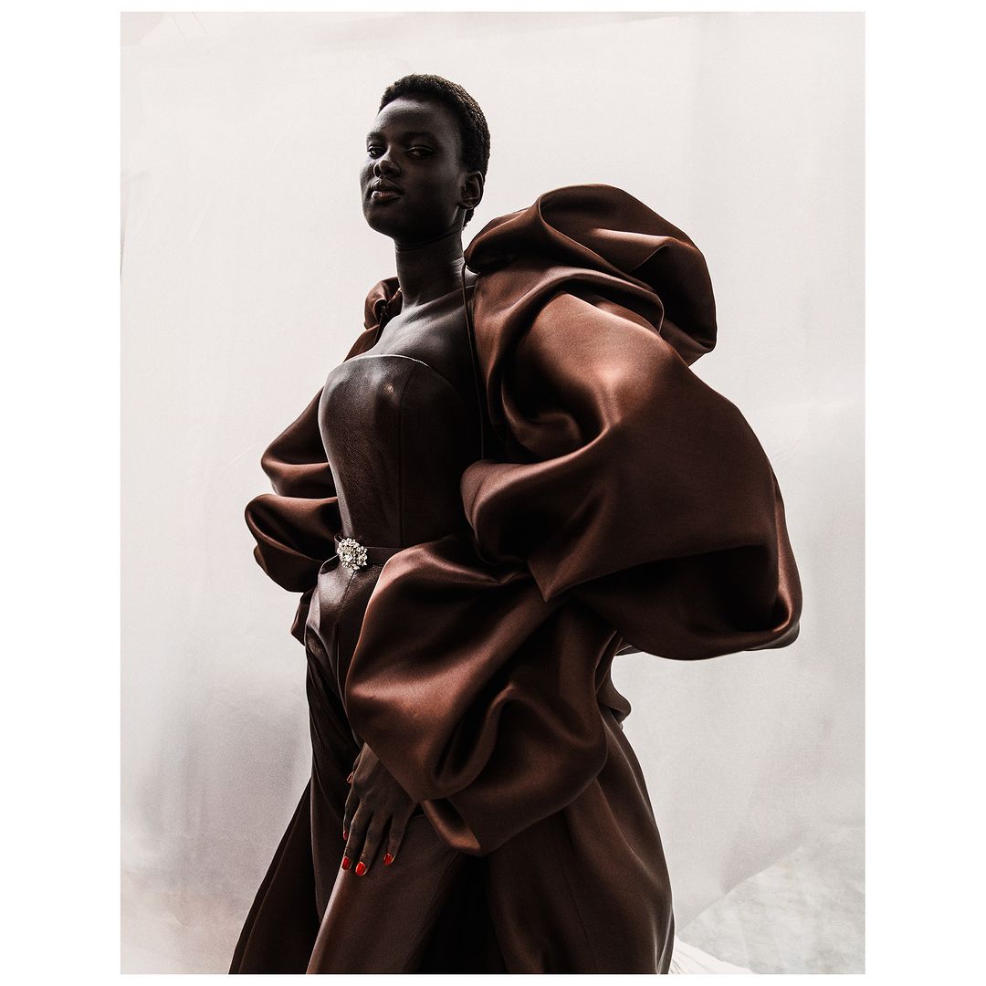 A woman with dark skin poses in a dramatic brown satin gown with large puffed sleeves against a neutral backdrop, fashion-forward and elegant.