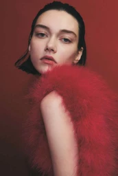 Close-up portrait of a woman with dark short hair wearing a fluffy red feather garment against a dark red backdrop.