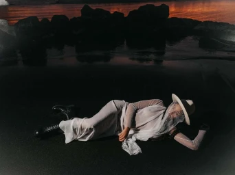 Woman wearing white dress and veiled hat lying on a dark sandy beach photographed by Filippo Fortis