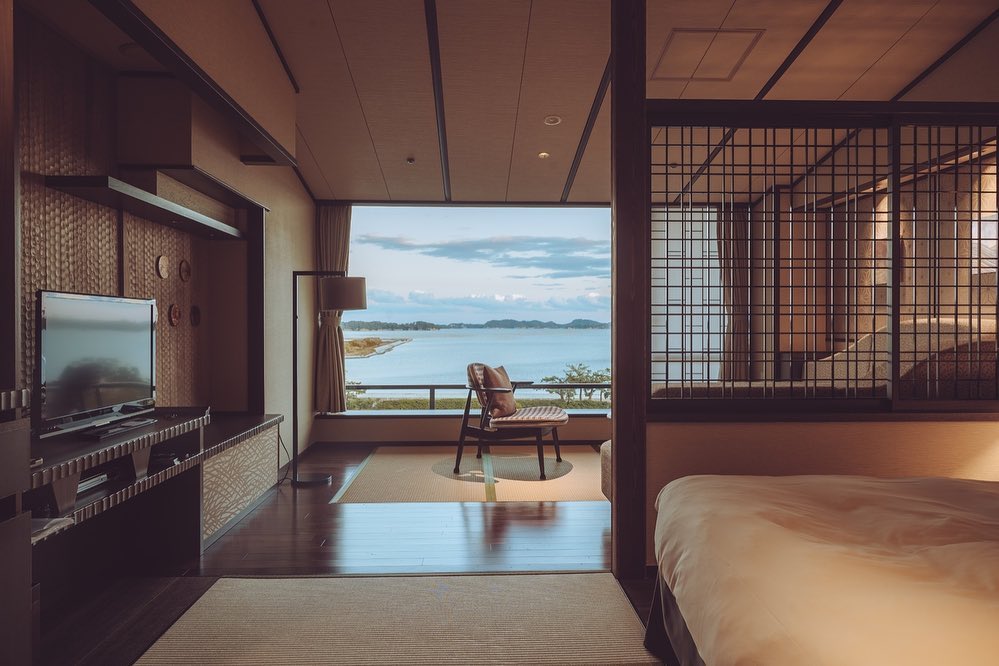 Interior of a hotel room with a view of the ocean; features include a television, a chair facing a window, and a bed. Warm, neutral tones.