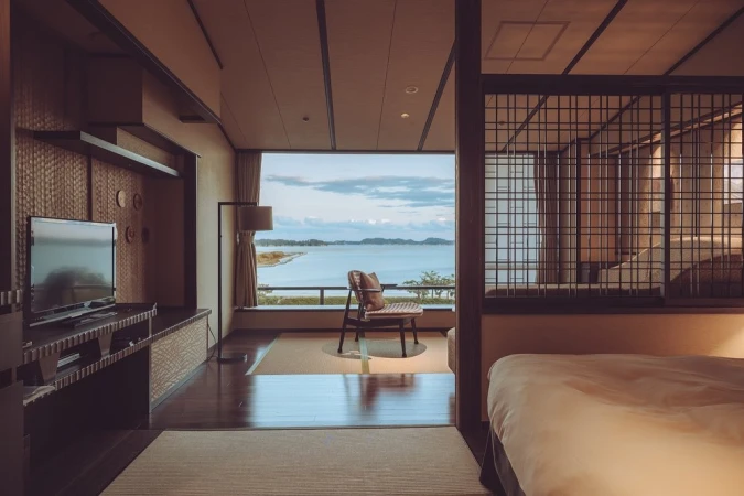 Interior of a hotel room with a view of the ocean; features include a television, a chair facing a window, and a bed. Warm, neutral tones.
