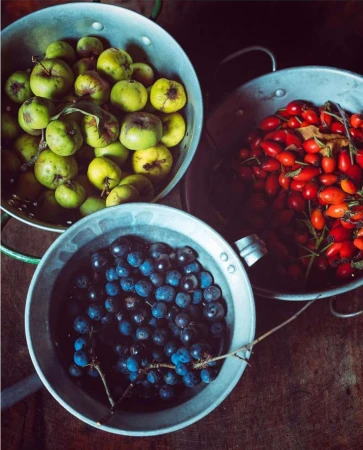 Overhead shot of three metal bowls filled with fresh green apples, red berries, and dark blue grapes on a rustic wood surface.