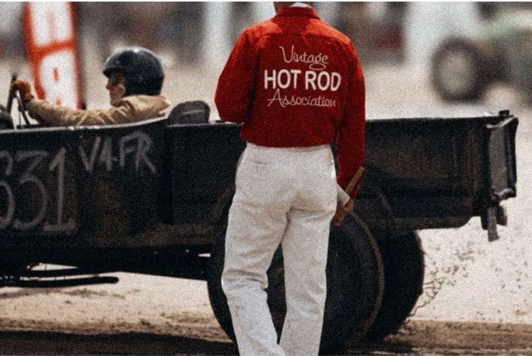 Hot rod racer wearing a helmet and goggles driving on a racetrack while an official stands nearby wearing a red jacket.