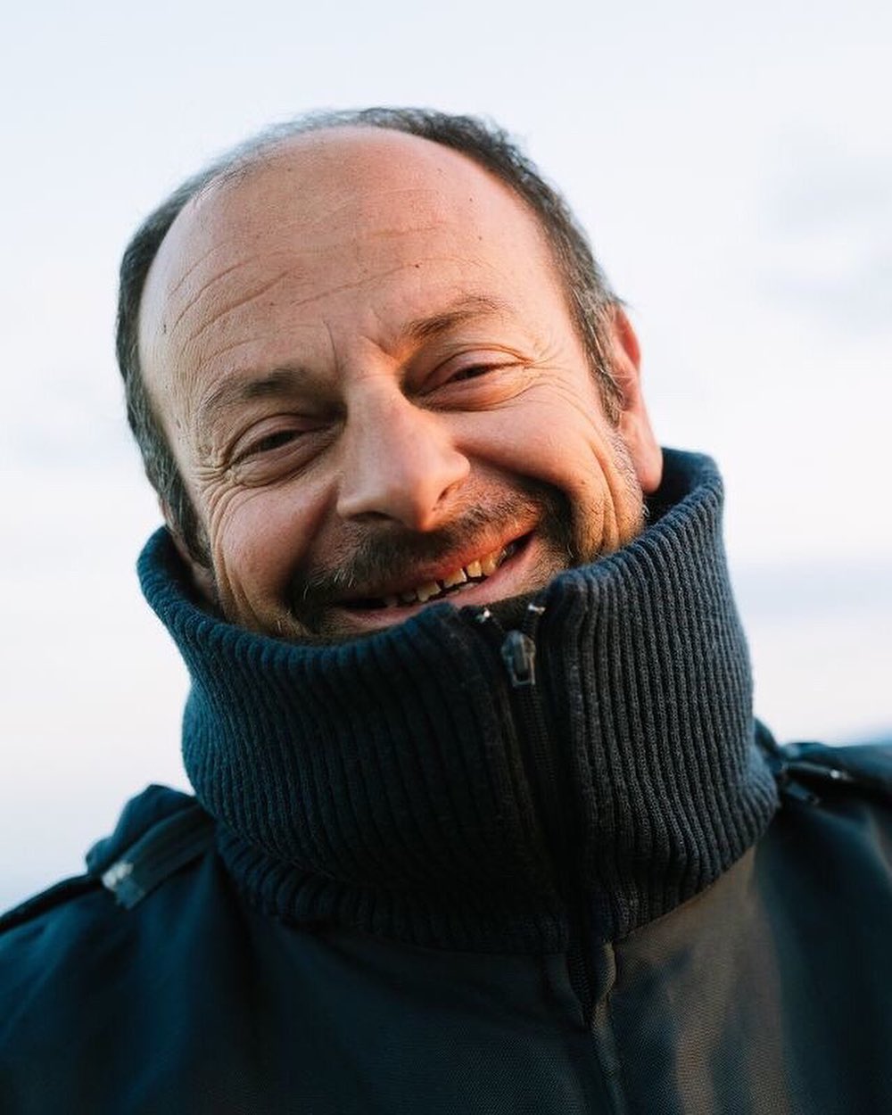 Close-up portrait of a smiling adult man with a shaved head outdoors, wearing a navy jacket with a high, ribbed collar.