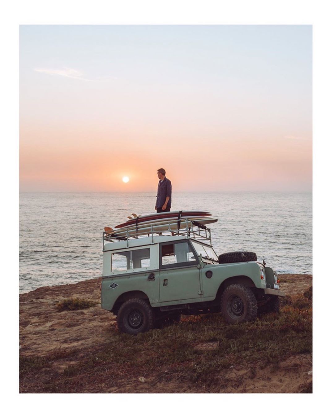 Man stands atop a classic Land Rover parked on a coastal cliff at sunset with surfboards, overlooking the ocean, creating a sense of adventure.