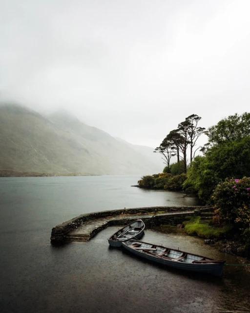 Landscape photo of two small rowboats docked at a stone pier on a lake, with forested hills in the background on a foggy day.