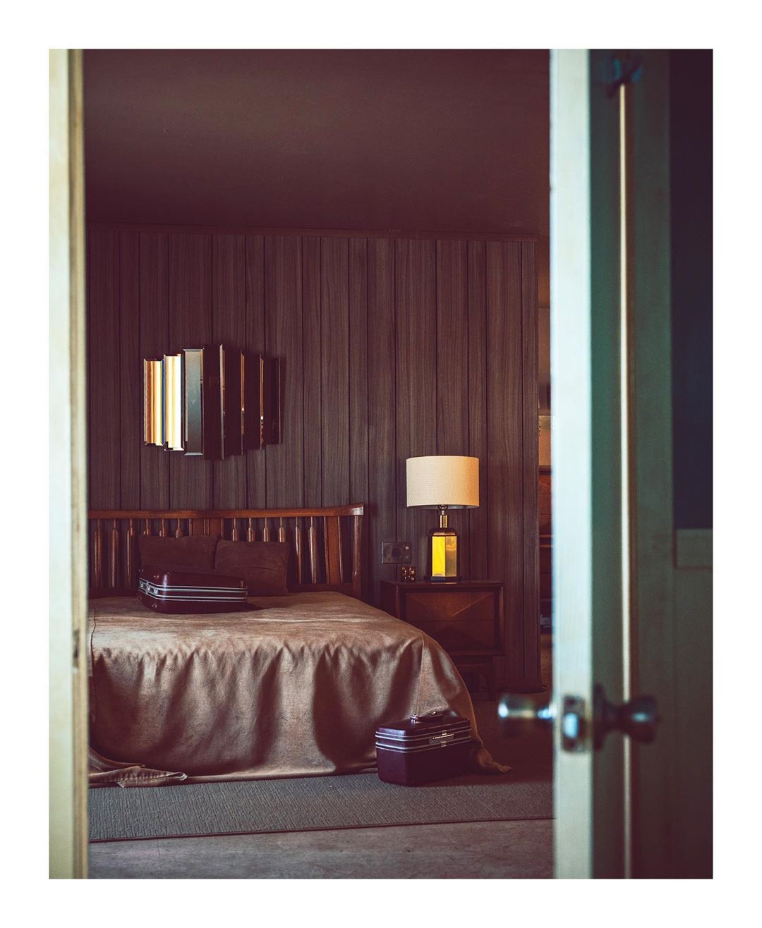 Hotel room interior with a bed, nightstand, lamp, luggage, and dark wood paneled walls as seen from doorway in a warm, inviting setting.