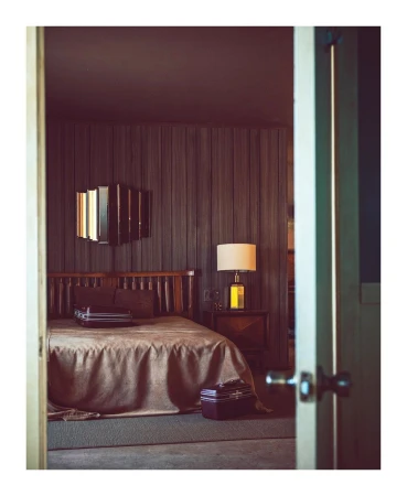 Hotel room interior with a bed, nightstand, lamp, luggage, and dark wood paneled walls as seen from doorway in a warm, inviting setting.