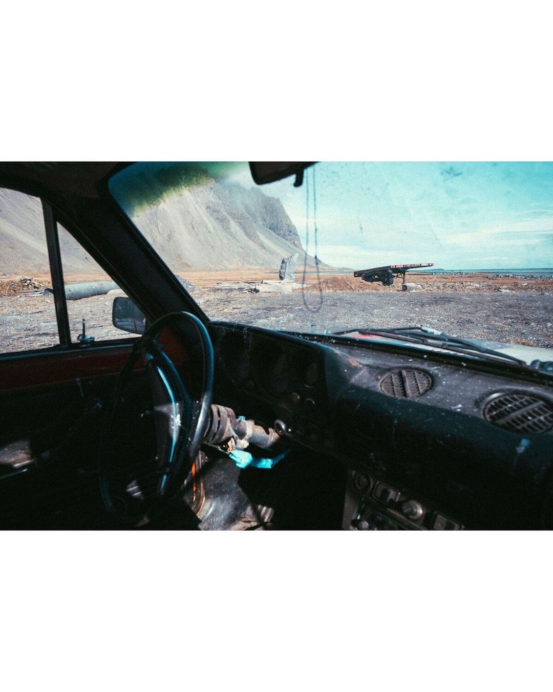 Interior view of a car parked on a beach with a mountain in the background on a sunny day showcasing the car's dashboard and steering wheel.