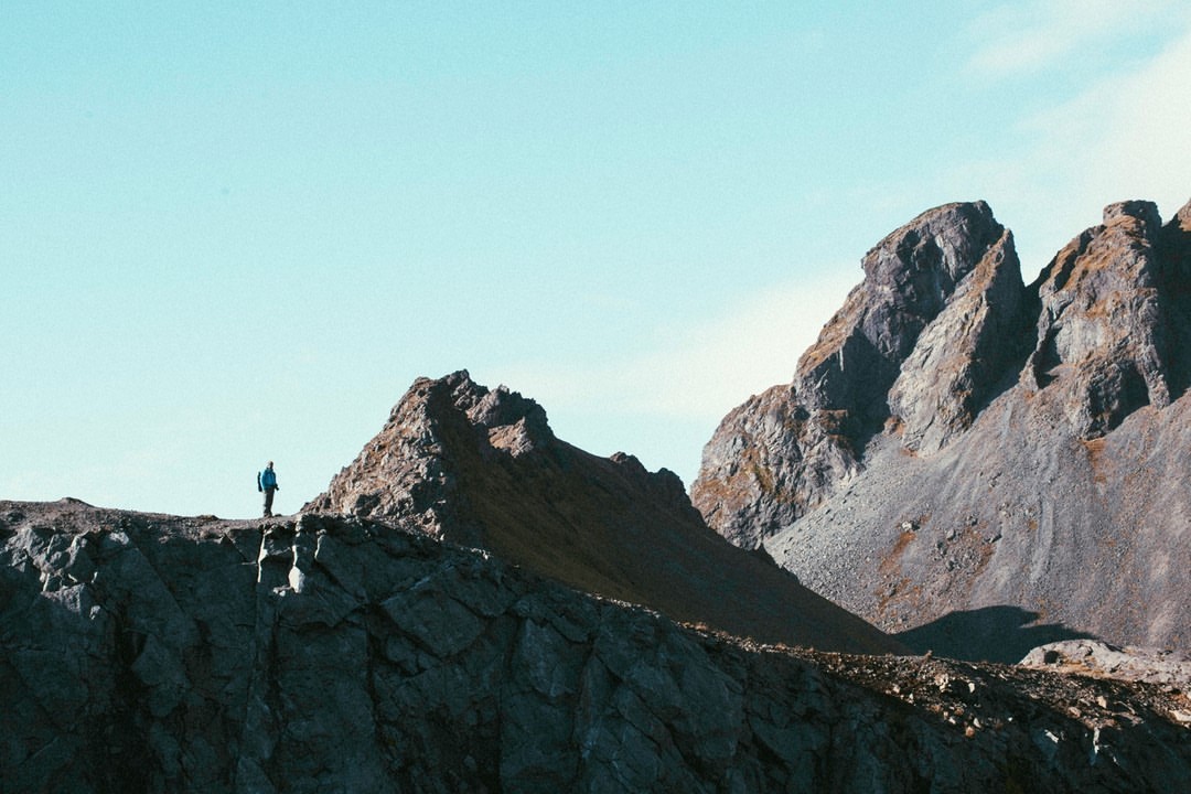A lone hiker in a blue jacket stands on a rocky mountain ridge against a pale sky, captured by photographer Finn Beales.