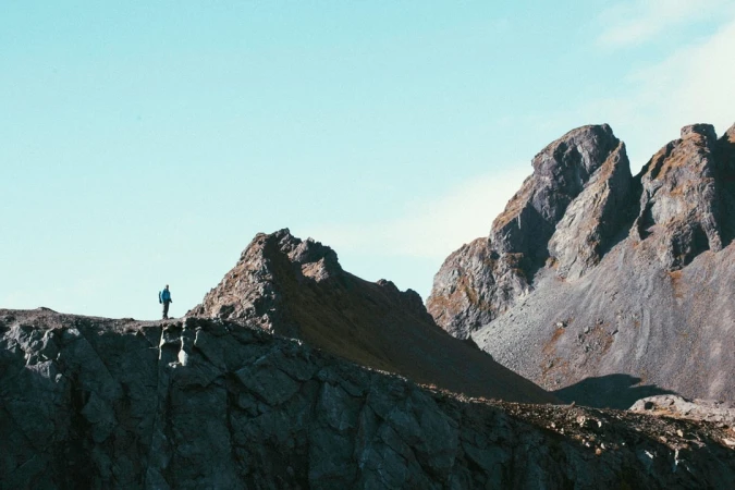 A lone hiker in a blue jacket stands on a rocky mountain ridge against a pale sky, captured by photographer Finn Beales.