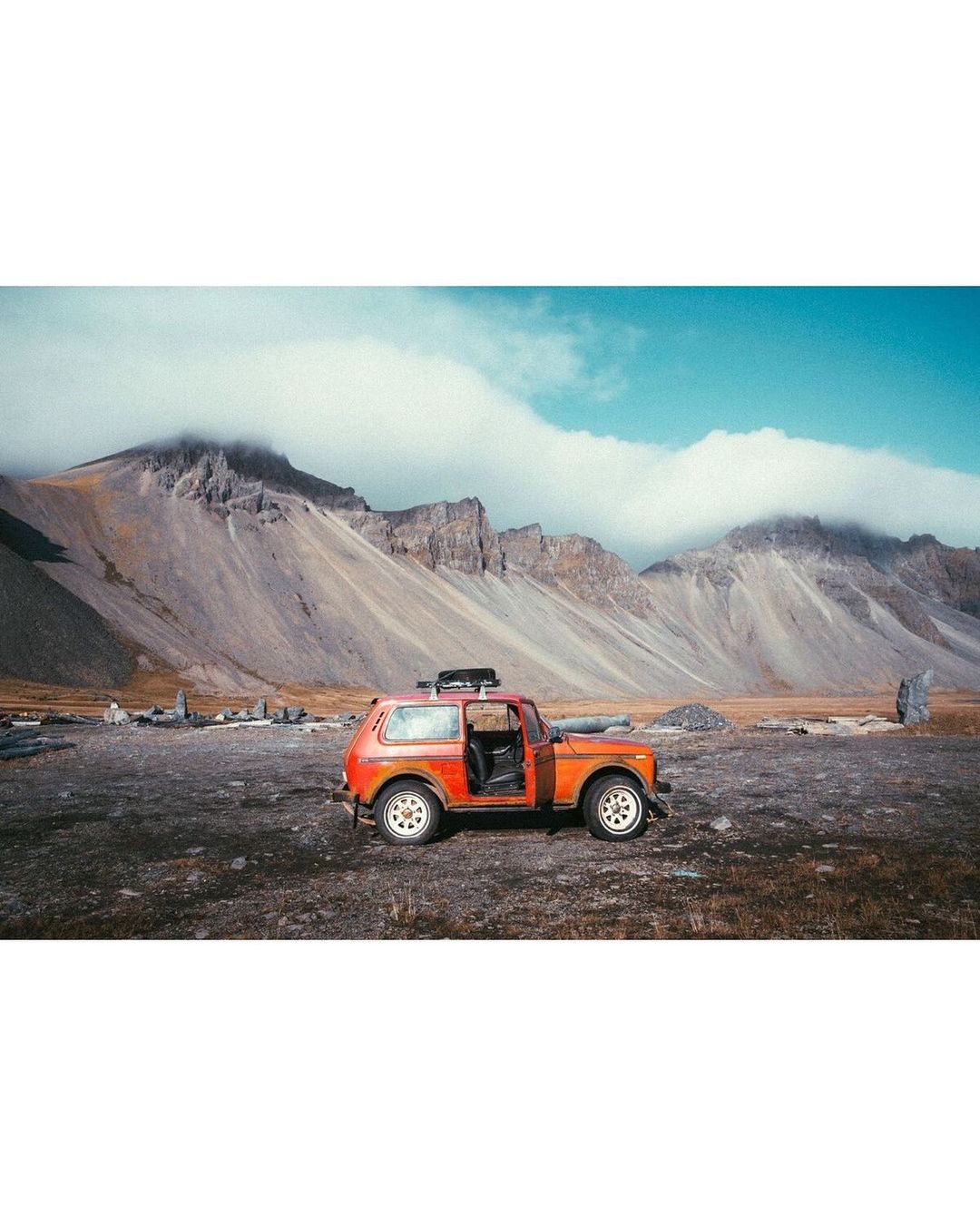 An orange off-road vehicle with its door open is parked on a rocky landscape in front of jagged mountains beneath a bright blue sky.