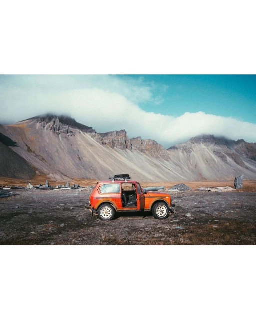 An orange off-road vehicle with its door open is parked on a rocky landscape in front of jagged mountains beneath a bright blue sky.