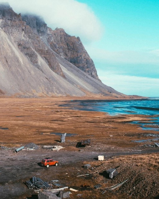 An orange car is parked on a dirt field in front of a tall, grey mountain near a body of water under a blue and cloudy sky.