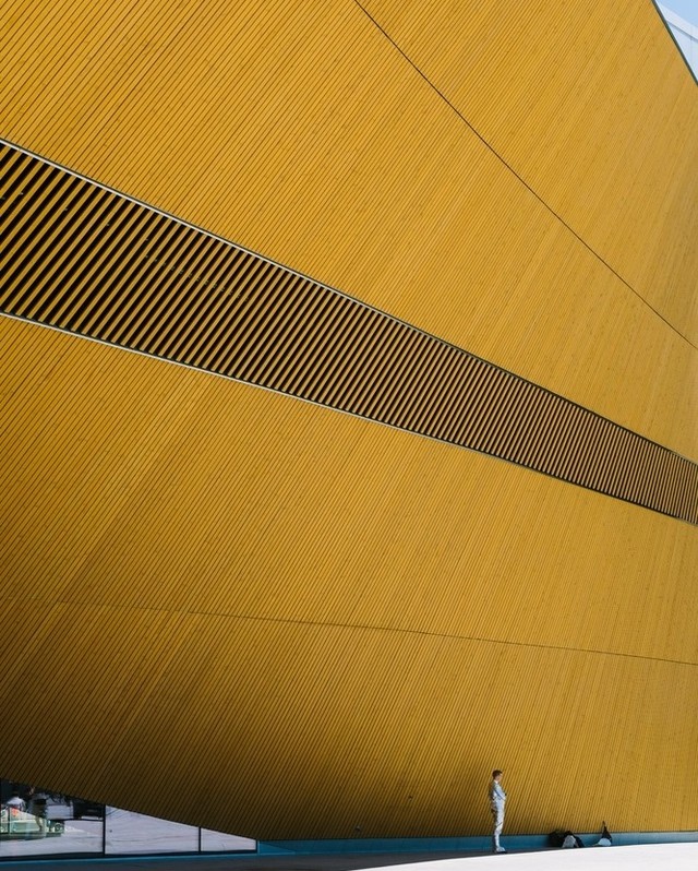 Person standing in front of a large, yellow building with black stripes, showcasing modern architecture and minimalist composition.