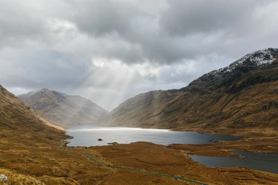 Scenic view of a lake surrounded by mountains under a cloudy sky, with light rays piercing through the clouds.