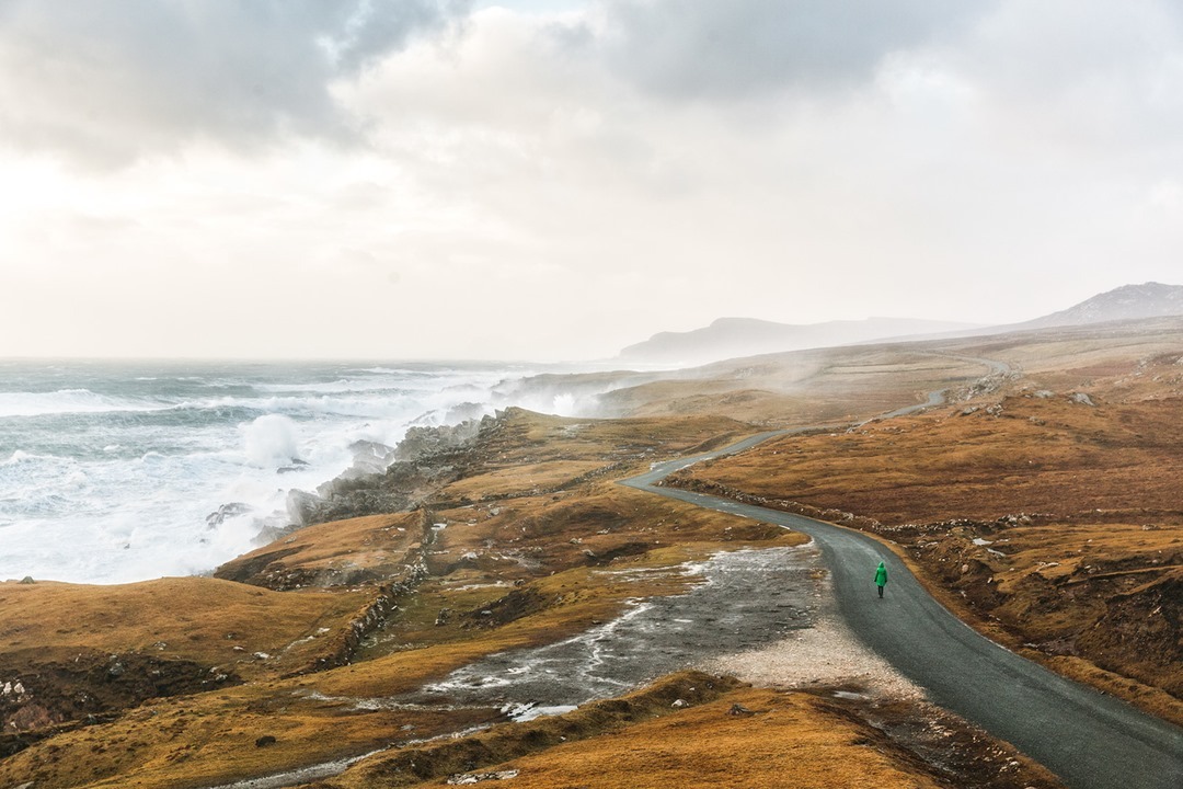 Adult in green coat walks along winding road, with rough coastline, crashing waves, and cloudy skies in a natural outdoor landscape.