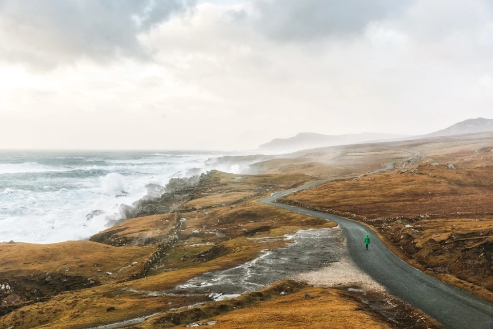 Adult in green coat walks along winding road, with rough coastline, crashing waves, and cloudy skies in a natural outdoor landscape.