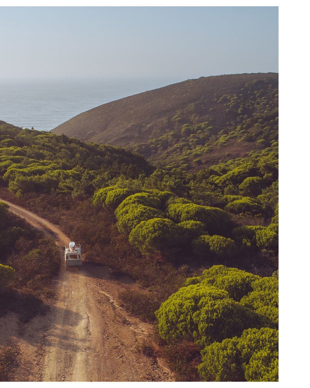 Overhead view of camper van driving on dirt road through green hills towards the ocean on a sunny day, under a clear blue sky.