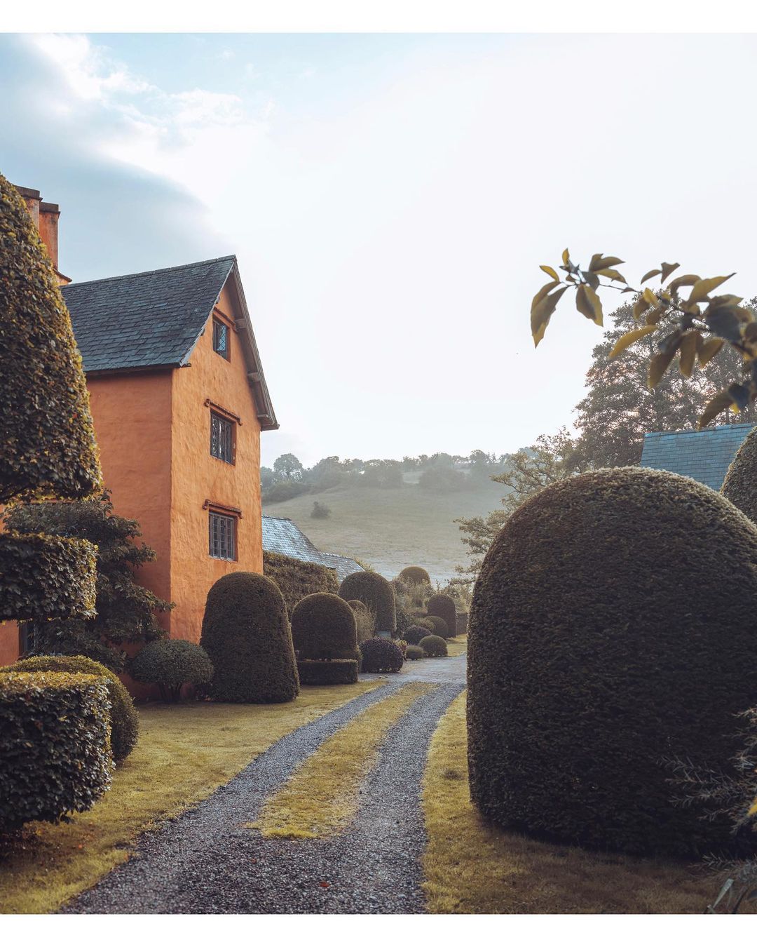 Exterior view of an orange brick house with a gray roof, manicured green topiaries, and a gravel driveway on an overcast day.