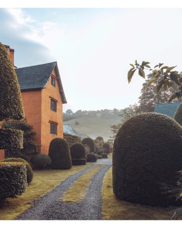 Exterior view of an orange brick house with a gray roof, manicured green topiaries, and a gravel driveway on an overcast day.