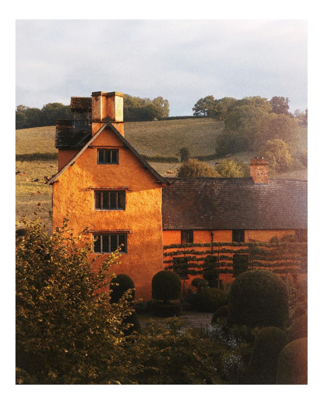 Exterior view of an orange country house with a dark tile roof and chimneys, surrounded by landscaped gardens and green fields.