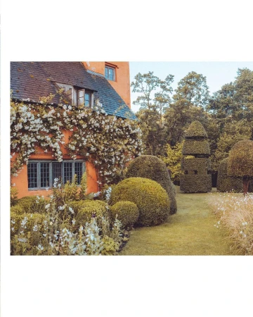 Exterior of a charming cottage house with peach-colored walls covered in white climbing roses, surrounded by a lush green garden with topiary.