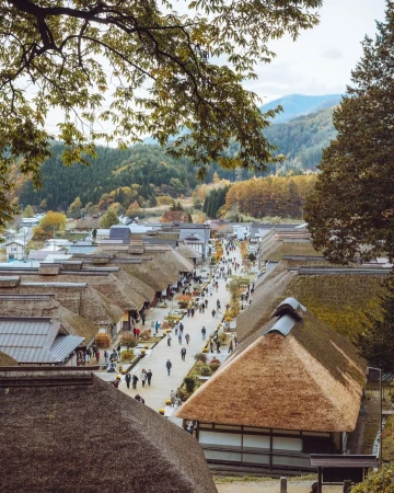 High angle view of a traditional Japanese village with thatched roofs, a road with people, and forested mountains in the background.