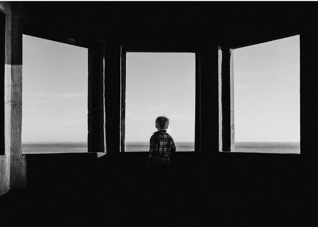 Monochrome photo of a child standing inside a concrete structure looking out at the ocean through multiple framed openings.
