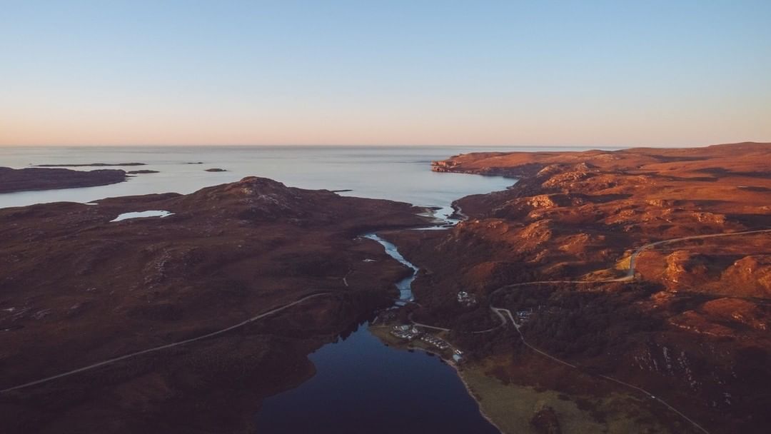 Aerial view of a rugged, brown landscape with a lake and ocean meeting under a clear sky. A road winds through the terrain.