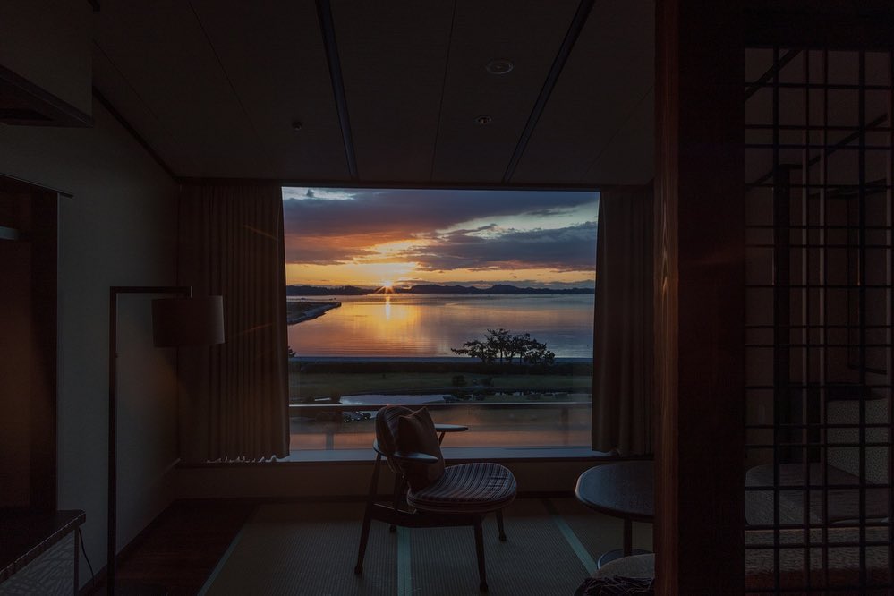Sunset view through a window in a room with a chair and floor lamp, offering a tranquil scene of water and distant land.