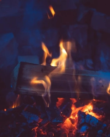 Burning wood and embers in a dark fireplace with orange and blue hues highlight the texture of the log and the glowing heat.