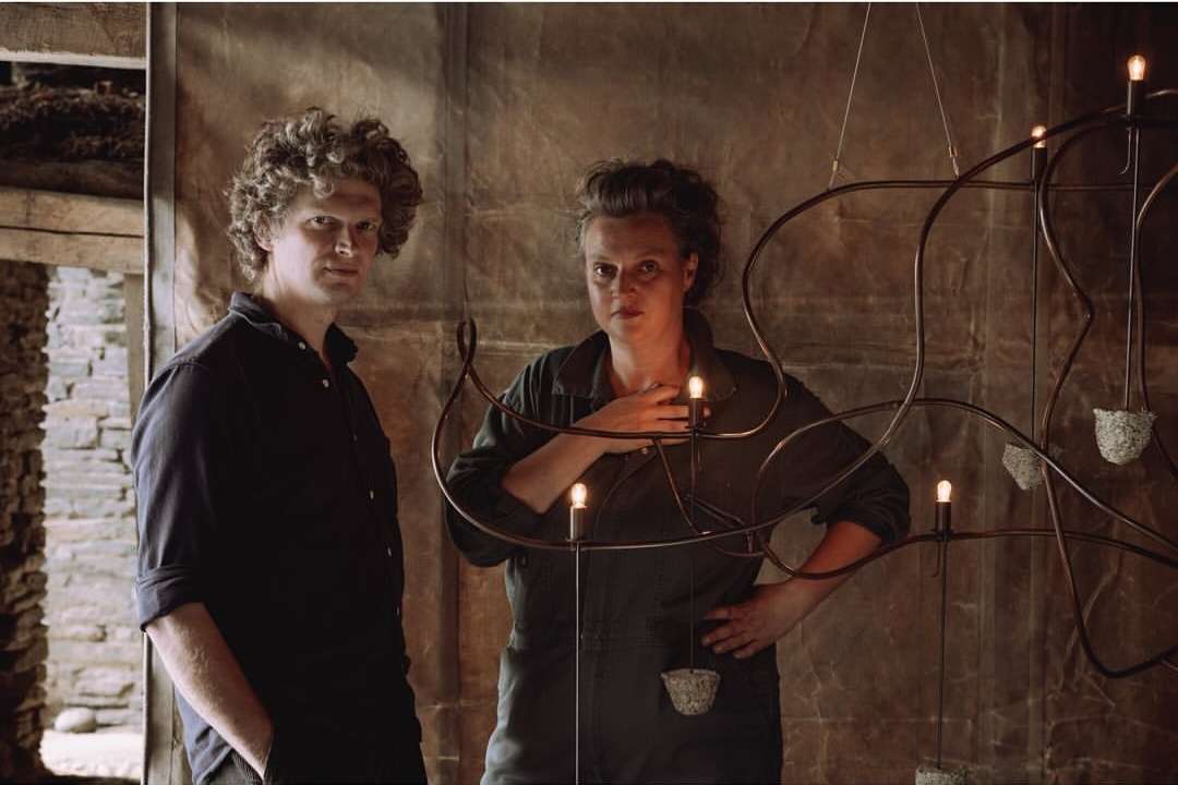 Man and woman stand in front of a unique chandelier with an industrial design in a rustic setting with earth-tone colors, indoor shot.