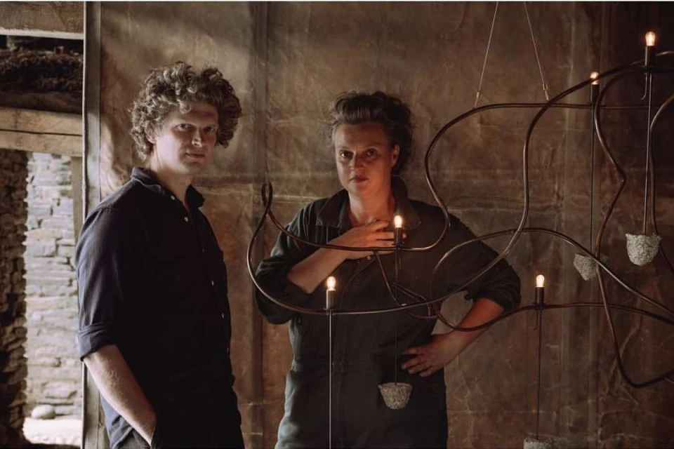 Man and woman stand in front of a unique chandelier with an industrial design in a rustic setting with earth-tone colors, indoor shot.