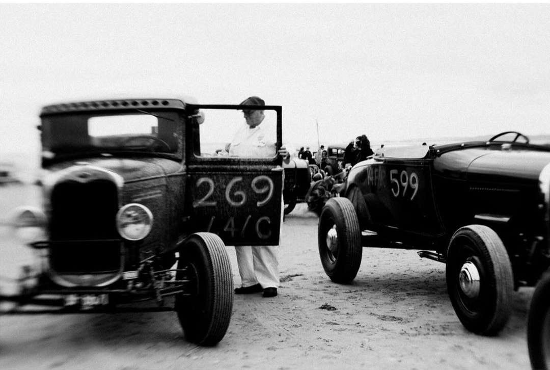Black and white shot of vintage cars lined up on a sandy surface, with a man standing by one of the car's open doors, racing number 269.