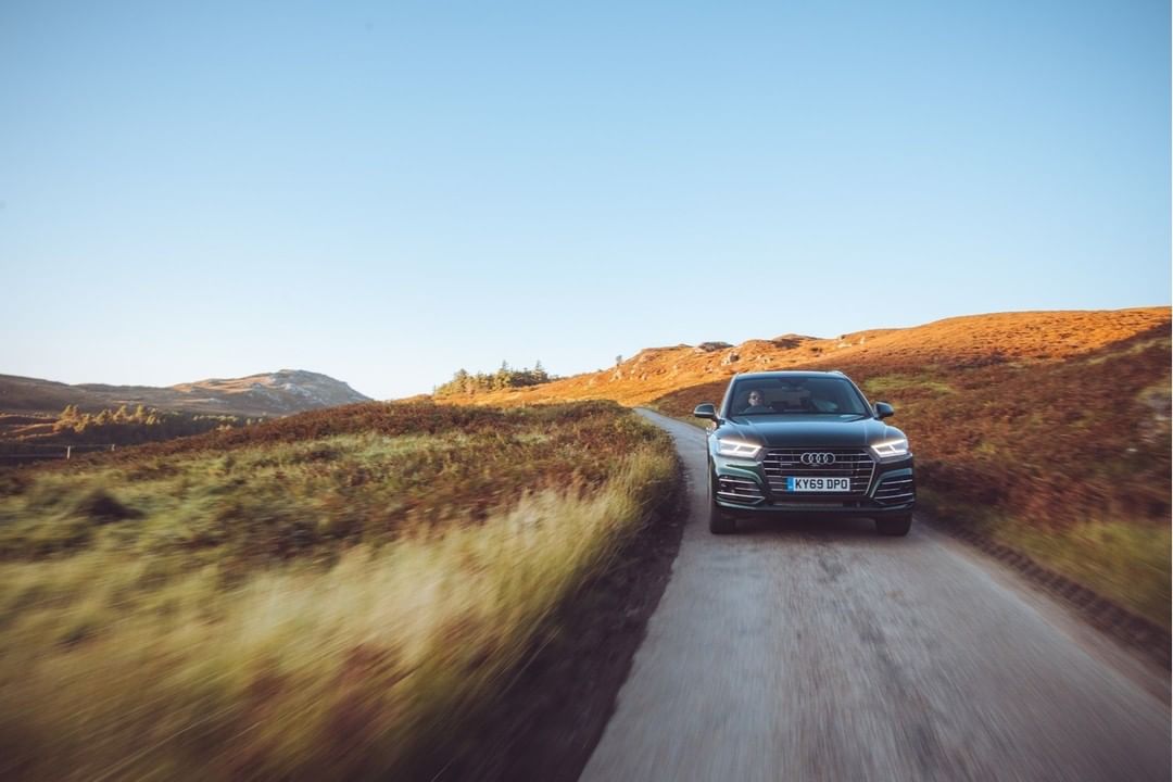 Green SUV drives down a rural road with grassy fields and hills on either side under a bright blue sky in the Scottish Highlands.