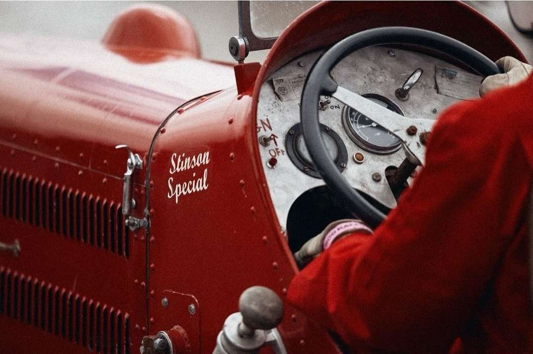 Close up of a vintage red Stinson Special race car with a driver wearing a red racing suit behind the steering wheel on a cloudy day.